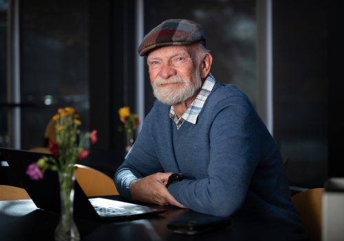 An older, smiling man sits at a desk with a laptop computer in front of him.