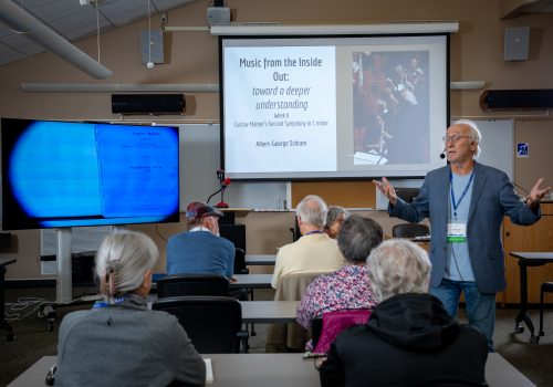 Older man speaking and gesturing with her arms, looking to her left. Older people sit at tables around him, listening attentively. | Image courtesy OLLI at UNC Asheville, photo Warner Photography.