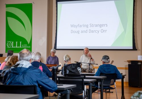 An older man and woman sit at the front of a classroom, speaking to an audience. A sign in the background identifies them as 