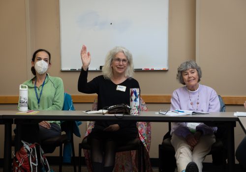 A group of three older woman sit at a table. The woman on the left wears a facemask, while the woman in the center raises her right hand.
