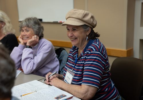 Two older women sit at a classroom table. The woman on the left smiles and rests her face on her hands, while the woman in the left has a pen in her right hand and an open notebook in front of her.