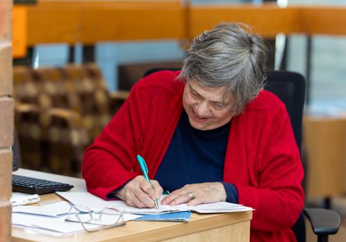 An older woman in a red sweater sits at a desk, writing on a stack of papers in front of her.