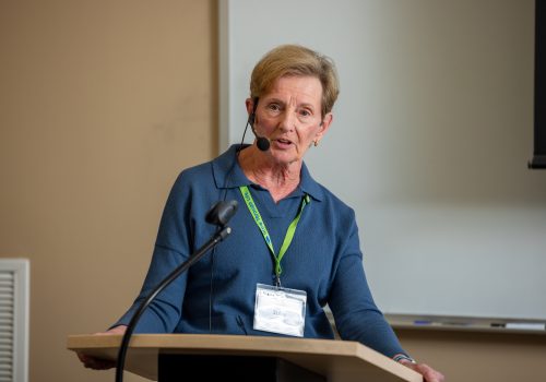 An older woman wearing a headset microphone speaks at a lectern.