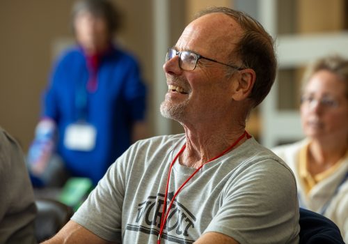An older man sits in a classroom with other people, smiling as he looks towards the front.