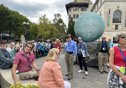 A group of older adults sits in a semi-circle in front of the Asheville Art Museum, listening to a tour guide through headphone devices.