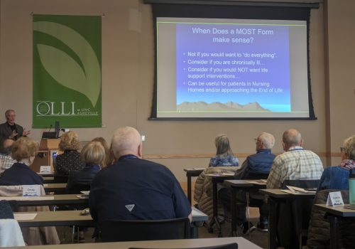 A group of older adults sit in a classroom, listening to a speaker seated to the left. A sign in the front of the room reads 